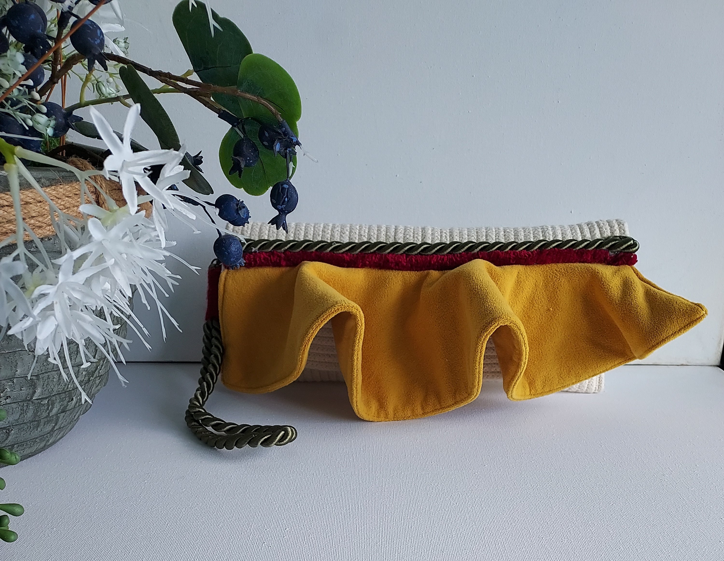 A mustard yellow fabric pouch with a ruffled edge, a dark brown braided cord, and a red inner lining, displayed on a white surface with decorative greenery.